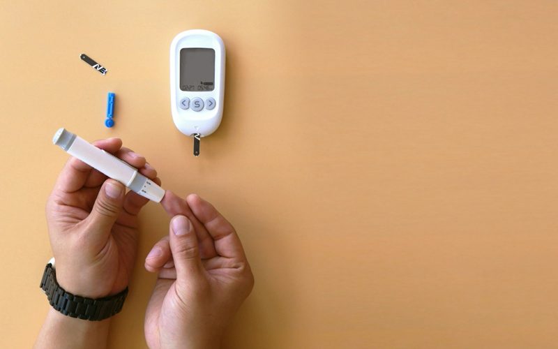 Mains tenant un lecteur de glycemie et une lancette sur fond orange